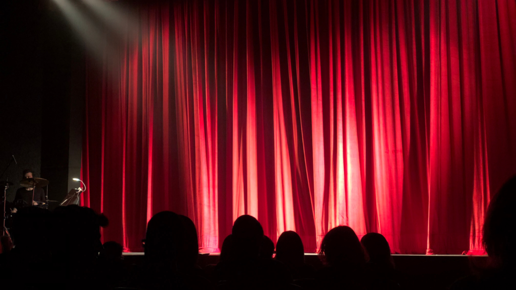 The silhouettes of an audience looking towards a theater stage with the red curtains drawn before an arts show begins