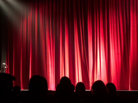 The silhouettes of an audience looking towards a theater stage with the red curtains drawn before an arts show begins